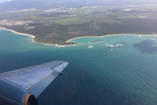 A plane wing out a plane window flying over the turquoise sea and white beaches of an island.