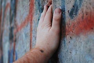 A close-up of a person’s hand resting gently on a worn, graffiti-covered concrete wall. The wall is cracked and layered with faded red and blue spray paint. The hand is steady and open, suggesting quiet presence or reflection.