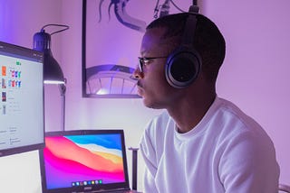 A Black man wearing headphones, in front of two monitors on a desk.