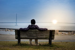 Person sitting on a bench in front of a lake looking at a suspension bridge with the sun rising