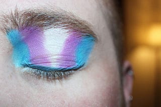 A close-up of a closed eye with the trans flag colours—blue, pink and white—painted on it with eyeshadow.