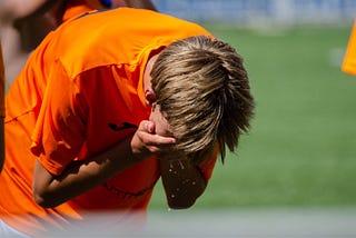 A child on a soccer field, with their head down, hands over their eyes in sadness