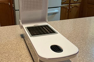 A Cuisinart countertop vegetable chopper with a clear container at the bottom and a hinged top lid. The lid has a textured pressing surface, and there’s a grid-style chopping blade insert in the center where vegetables are pushed through to create diced pieces directly into the container below.