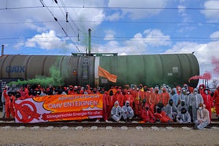 Aktivisti blockieren das Raffineriegleis vor einem Tankerwagen. Verkleidet in orangen und weissen Anzügen mit einem Transpi worauf „Klimazerstörin OMV enteignen! Klimagerechte Energie für alle!’’