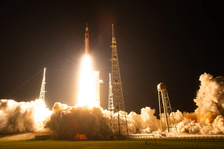 A rocket launch at night. The glow from the rocket illuminates the launchpad and smoke surrounding it as it begins to fly.