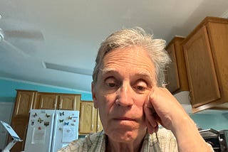 An older man with gray hair sits in a kitchen, resting his face on his hand and looking doubtfully at the camera.
