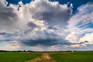An image of growing cumulonimbus clouds over a flat, grassy midwestern field.