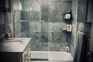 Image shows a bathroom, focused on the dry tub, surrounded by carrera marble tiles, with a moody blue tone on the image.