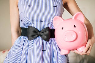 A young woman in a blue pinstriped dress with a black bow belt holding a pink piggy coin jar at her hip.