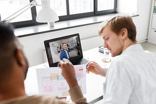 Two designers reviewing a paper prototype with sticky notes while conducting a remote customer interview with a participant visible on the laptop screen
