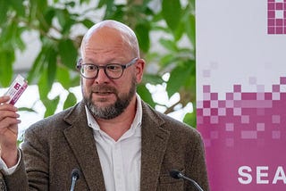 Chief Digital Officer for London, Theo Blackwell, giving a speed to an audience of data experts at City Hall in London. He’s holding a sticker that says: ‘I search city data in one place at Data for London libary — dfl.london.gov.uk/library’