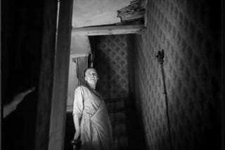 A still from Housing Problems (1935) .A woman stands at the top of crooked stairs in her house. A hole in the ceiling is visible above.