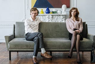 A couple having issues in their relationship, sitting on a gray couch with their legs crossed, separated from each other.