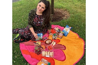 Brunette millennial in a dress sits on a round towel covered in snacks sometime in 2019.
