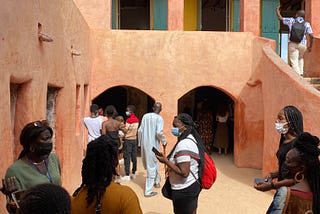 A group of people outside the House of Slaves (Maison des Esclaves), a red building made of clay.