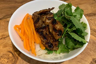 Grilled pork, rice noodles, pickled carrots and herbs served in a white bowl on a wooden table.