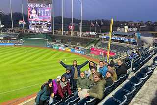 People from the Monarch team and other Biocuration Conference attendees look back towards the camera while siting in the baseball stadium with the field behind them.