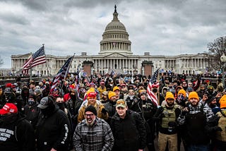 U.S. capitol insurrectionists wave American flags dressed in military gear, gather in front of the U.S. capitol building. One person in front is screaming with rage.