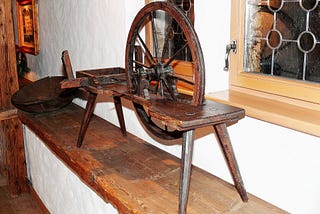A photo of a wooden spinning wheel sitting on a shelf by a window.