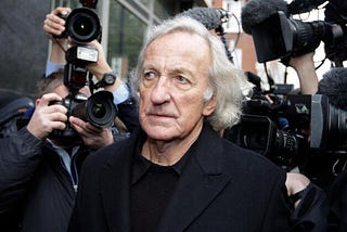 Older man with grey hair surrounded by media cameras.