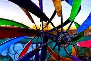 A closeup of a windmill with many colors in front of a sunset and a fence covered in graffiti writing something indeciferable.