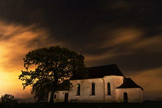 Under a dark starry sky, a white concrete haunted house with a black roof rests next to a tree.