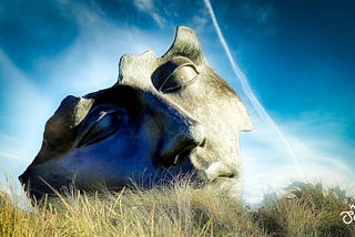 Blue Sky photo of a broken mask sculpture in a grassy field.