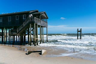 Photo of a home on stilts in the water next to a few pilings in the water from a previous home that was washed away