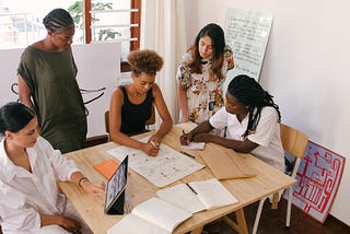 Group of team members working together to brainstorm stakeholders and team process. They are sitting and standing around a table, some with paper and pens and others with computers.