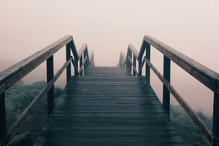 A dark wooden slatted walkway with wooden sides rails, laid over mossy rocks and leading down into a fog that obscures everything in front of it.