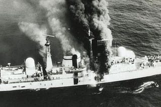 A black and white image showing HMS Sheffield on fire after the attack. Crewmbers can be seen at the bow and stern.