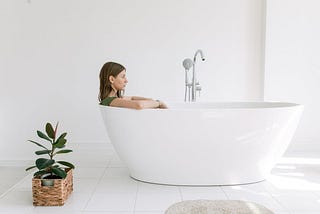 A woman wearing a shirt sits in a white bathtub, staring blankly ahead. There is a plant in a wooden basket near the tub.