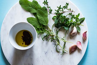 Several plants & leaves lie next to a cup filled with oil. All are resting on a circular white marble stone on a blue table