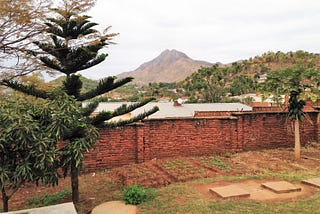 In the distance there is a brown mountain with a tree covered hill in front. In the foreground there is a brick wall with a garden in front of it, and some trees framing the view off to the left.