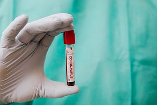 White gloved hand holding a clear vial labeled “coronavirus” with a red top. In the background is part of a blue disposable medical gown