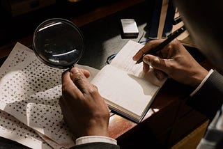 A man’s hand holding a magnifying glass in his left hand and writing down notes with his right hand in a notebook. He is looking at some coded papers.