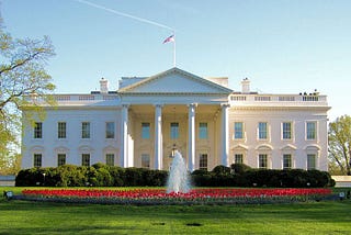 Photo of the White House from the North on a sunny day. There is a fountain surrounded by red flowers in the foreground.