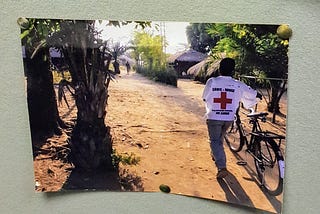 Image of a photo pinned to a noticeboard. The photo shows a man in red cross uniform arriving at a rural village.