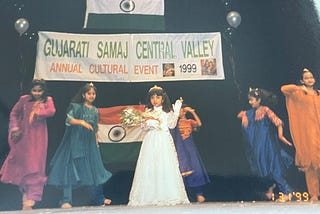 A 5-year-old Indian girl wears a white dress, tiara, and sash as she stands onstage. Surrounding her are other Indian girls, dancing in salwaar kameez outfits of differing colors.