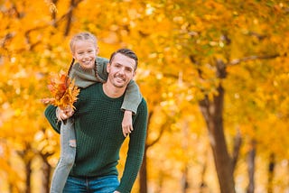 Father carries daughter through autumn trees. She holds yellow orange leaves.