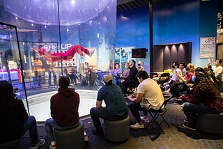 Students sitting around an air tunnel and watching an indoor skydiver take flight.
