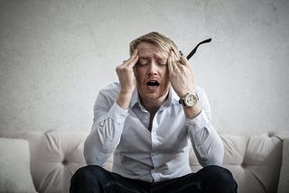 White, blond- haired man with a white button up shirt, black slacks and a watch on his left wrist sits on a couch with his hands on his temples looking distressed and tired. He signifies a chronic energy supplier.