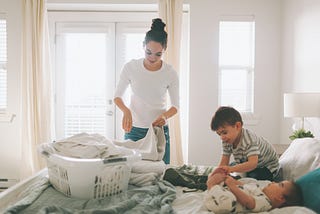 A mother folding laundry with her children in a bright, clean bedroom.