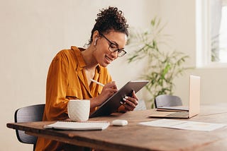 Photo of a woman in an orange shirt sitting at a table and taking notes on a tablet.