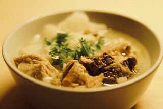 A white ceramic bowl filled with mutton soup, showing pieces of lamb meat, green herbs, potato cubes, and broth