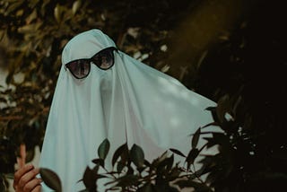 A woman with sunglasses on wearing a ghost costume in a bush.