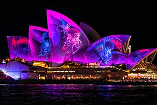 An image of the Sydney Opera House illuminated with purple lights and at night.