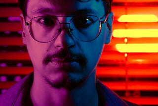 A portrait of the author looking straight at the camera. The left side of his face is lit in pink while the right side is lit in blue. In the background, a circular orange light pierces through horizontal blinds.