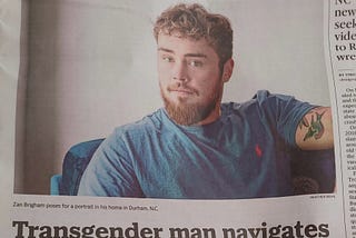 A photograph of a News&Observer newspaper shows a white trans man in North Carolina. He is blond, has a full beard, is wearing a blue t-shirt, and has a tattoo on his arm. The article in the newspaper is titled “Transgender man navigates anti-LGBTQ climate in NC”.