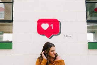 A lady standing against a wall with a red speech bubble that has a single heart and the number one next to it above her head. The lady is turning her head to look away from the camera and looks uneasy, with her hand curling her hair behind one ear.